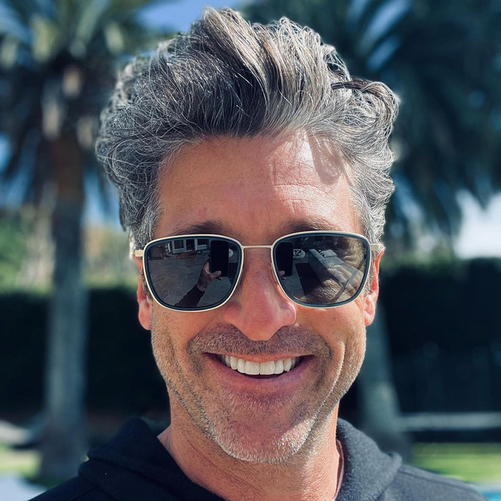 Closeup selfie of Patrick Dempsey smiling wearing black, his phone reflected in his glasses.